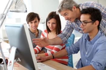 Students sitting around computer with instructor standing above pointing at screen